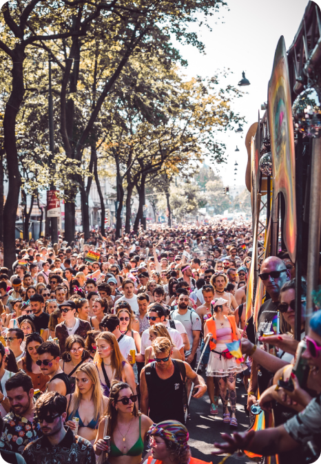 Crowd of diverse individuals joyfully participating in the Vienna Pride 2022 parade, marching down a tree-lined street. Participants are seen wearing colorful clothing and accessories, embodying the spirit of Pride with smiles and raised rainbow flags, celebrating LGBTQ+ rights and visibility in a sunny, vibrant setting.