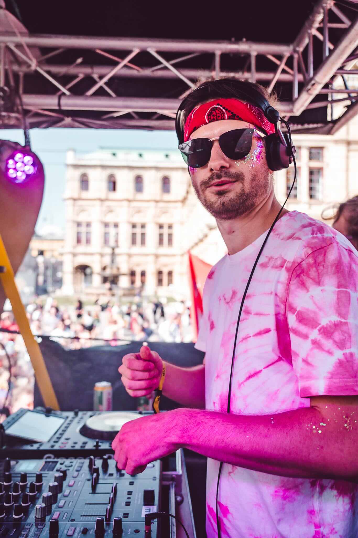 Dynamic image of a DJ at a live event, wearing a red bandana and sunglasses, actively adjusting controls on a DJ mixer. He is dressed in a pink tie-dye shirt, with a touch of glitter on his face, capturing the lively and festive atmosphere of the scene. The backdrop features the historical architecture of a city square, suggesting a vibrant outdoor music festival setting.