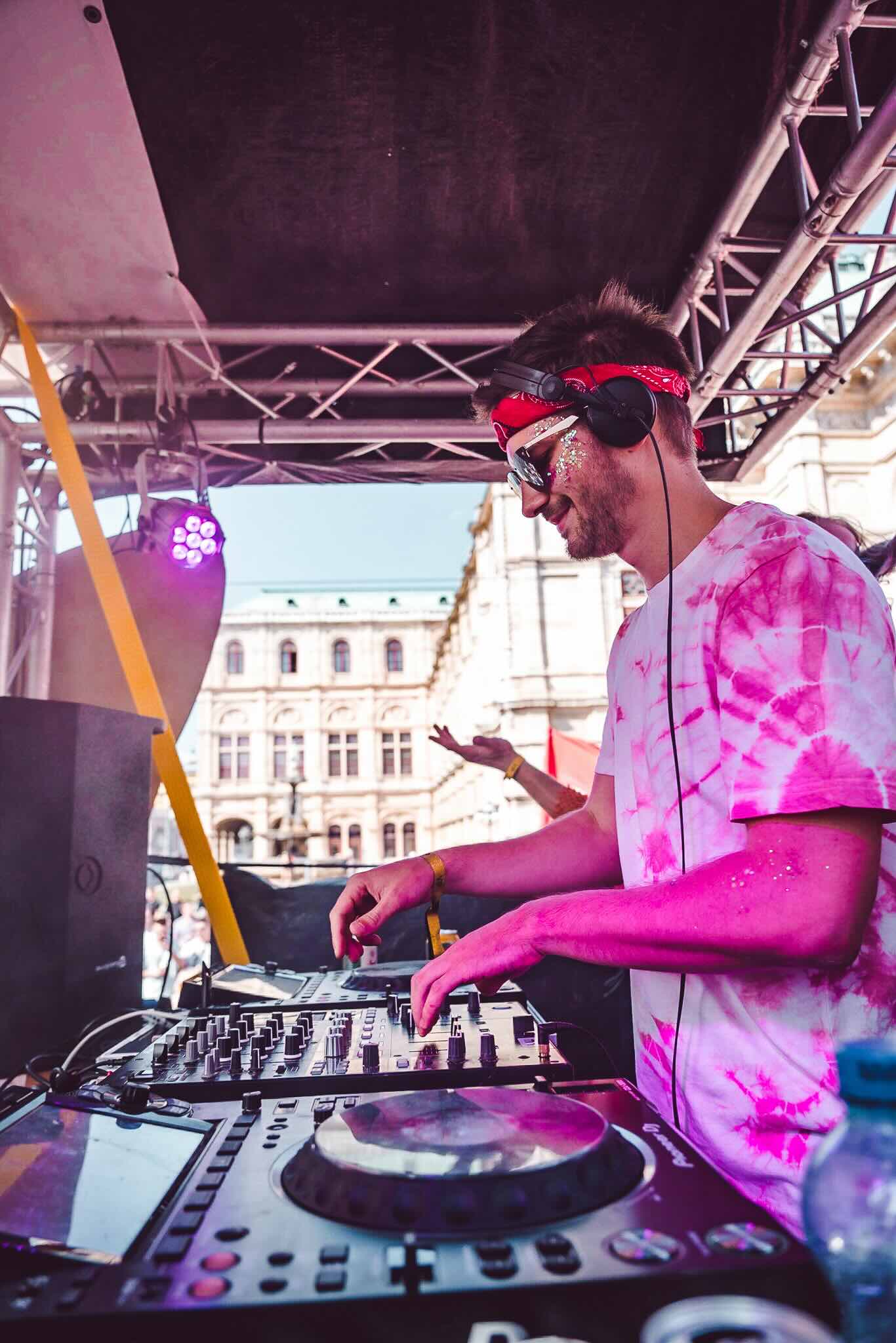 Dynamic image of a DJ at a live event, wearing a red bandana and sunglasses, actively adjusting controls on a DJ mixer. He is dressed in a pink tie-dye shirt, with a touch of glitter on his face, capturing the lively and festive atmosphere of the scene. The backdrop features the historical architecture of a city square, suggesting a vibrant outdoor music festival setting.
