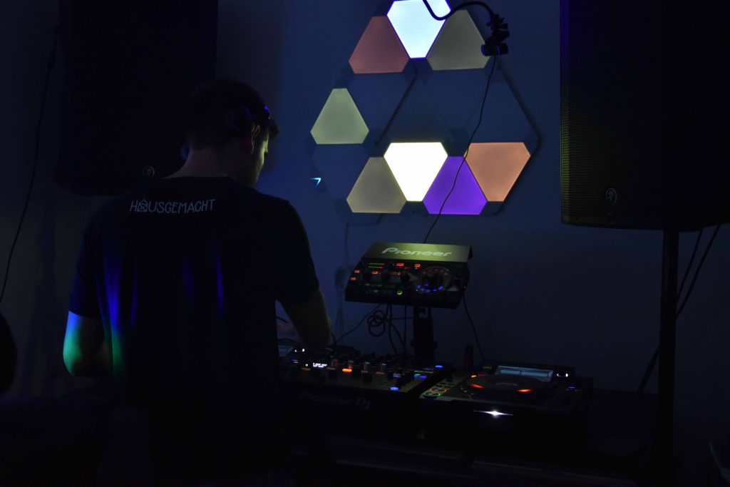 DJ in silhouette against an illuminated geometric wall installation, working a Pioneer DJ deck in a dark room. The hexagonal shapes on the wall emit soft, multicolored lights, casting a cool ambiance. The back of the DJ's shirt reads 'HAUSGEMACHT'.