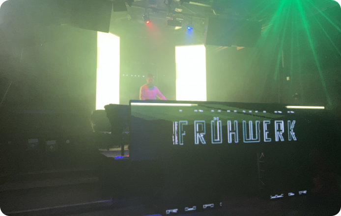 Silhouette of a DJ performing on stage at Fluc Wanne with bright vertical light panels on either side, atmospheric stage lighting with green beams, and the text 'Frühwerk' illuminated on the front of the DJ booth, creating a dynamic club atmosphere.