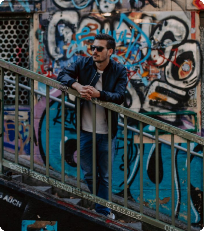 Man in a casual pose leaning on a railing in front of a vibrant graffiti wall, wearing a dark leather jacket, jeans, and sunglasses, embodying a cool and urban vibe.