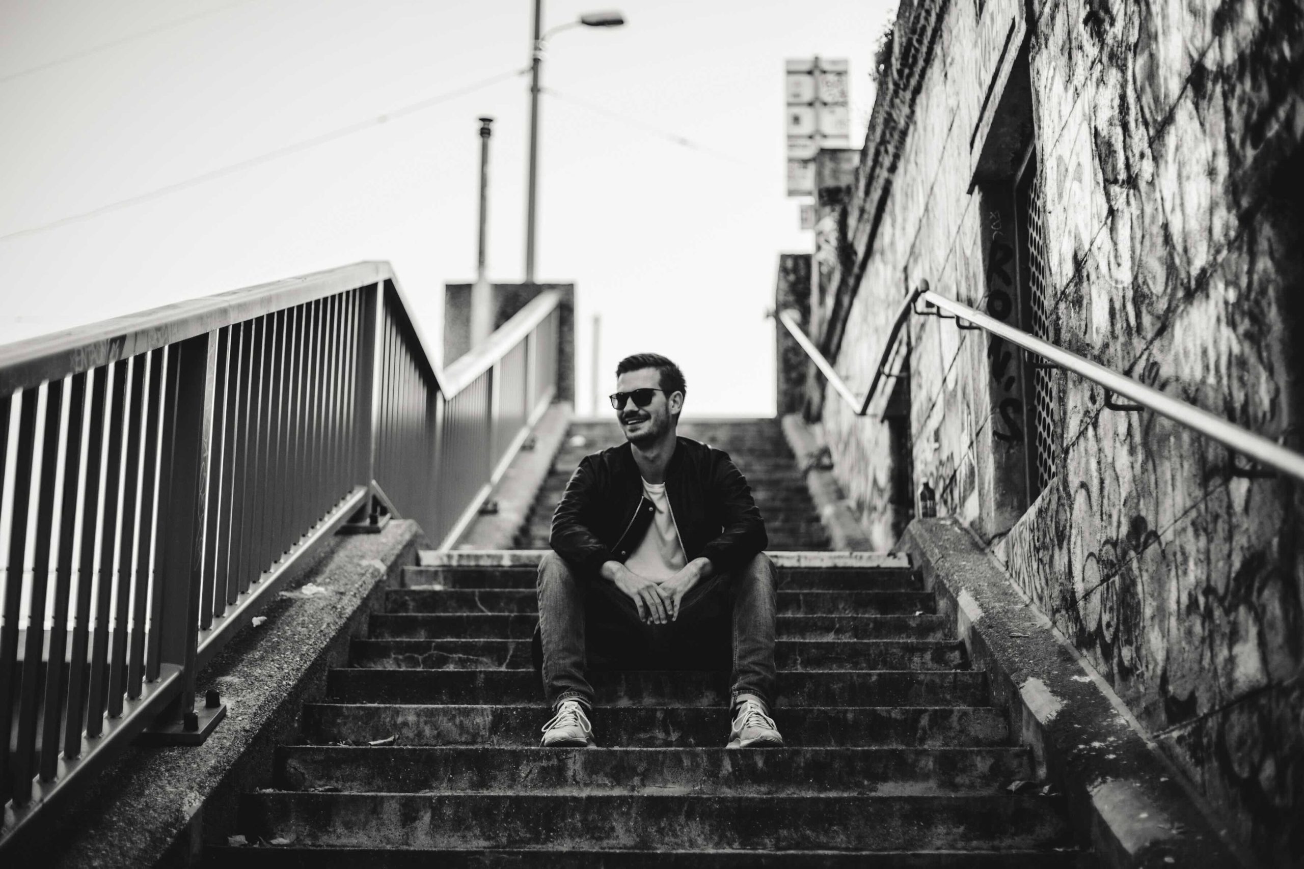 Man in a casual pose leaning on a railing in front of a vibrant graffiti wall, wearing a dark leather jacket, jeans, and sunglasses, embodying a cool and urban vibe.